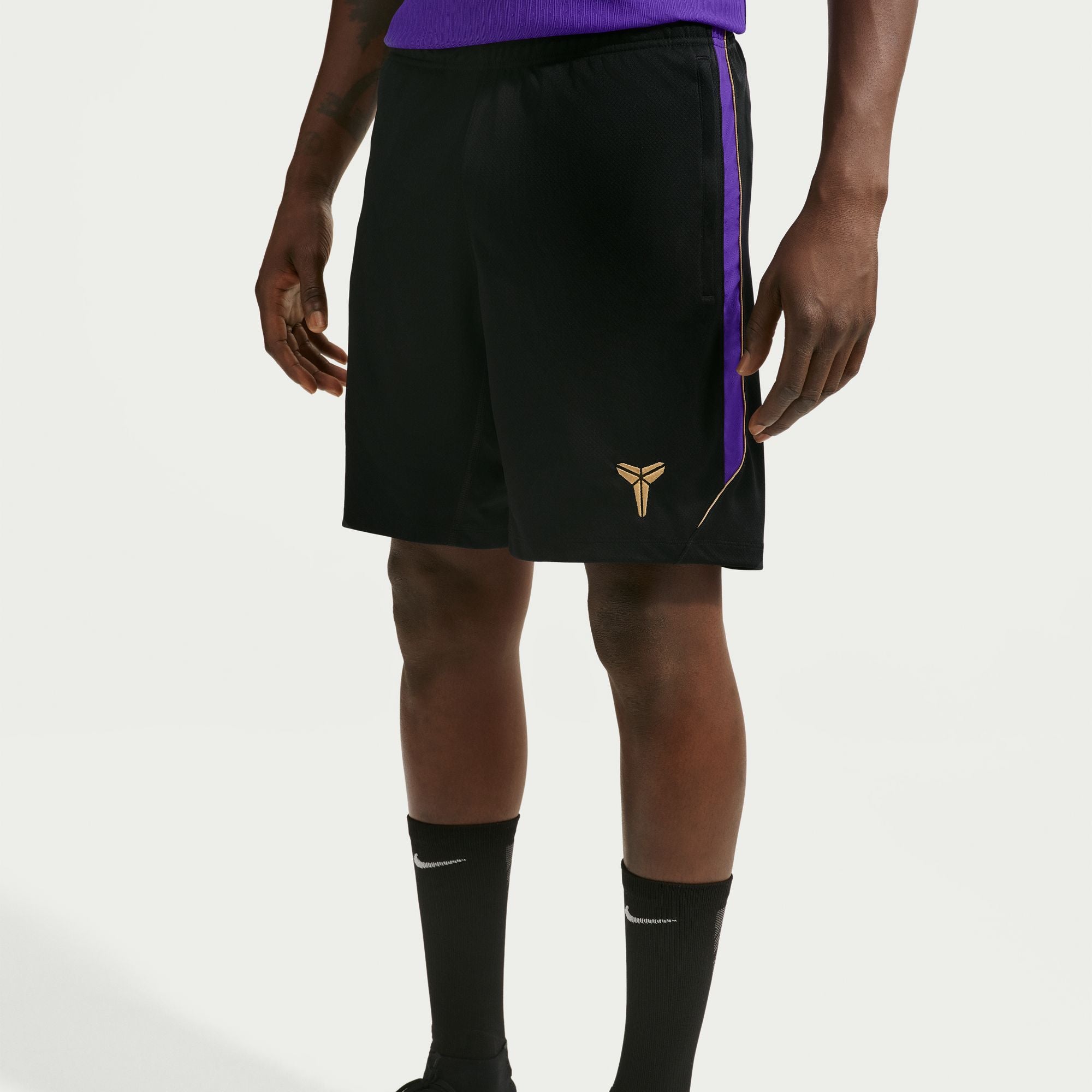 Black athletic shorts with a purple side stripe and a gold Kobe Bryant logo, worn by a model with black socks against a light gray background.
