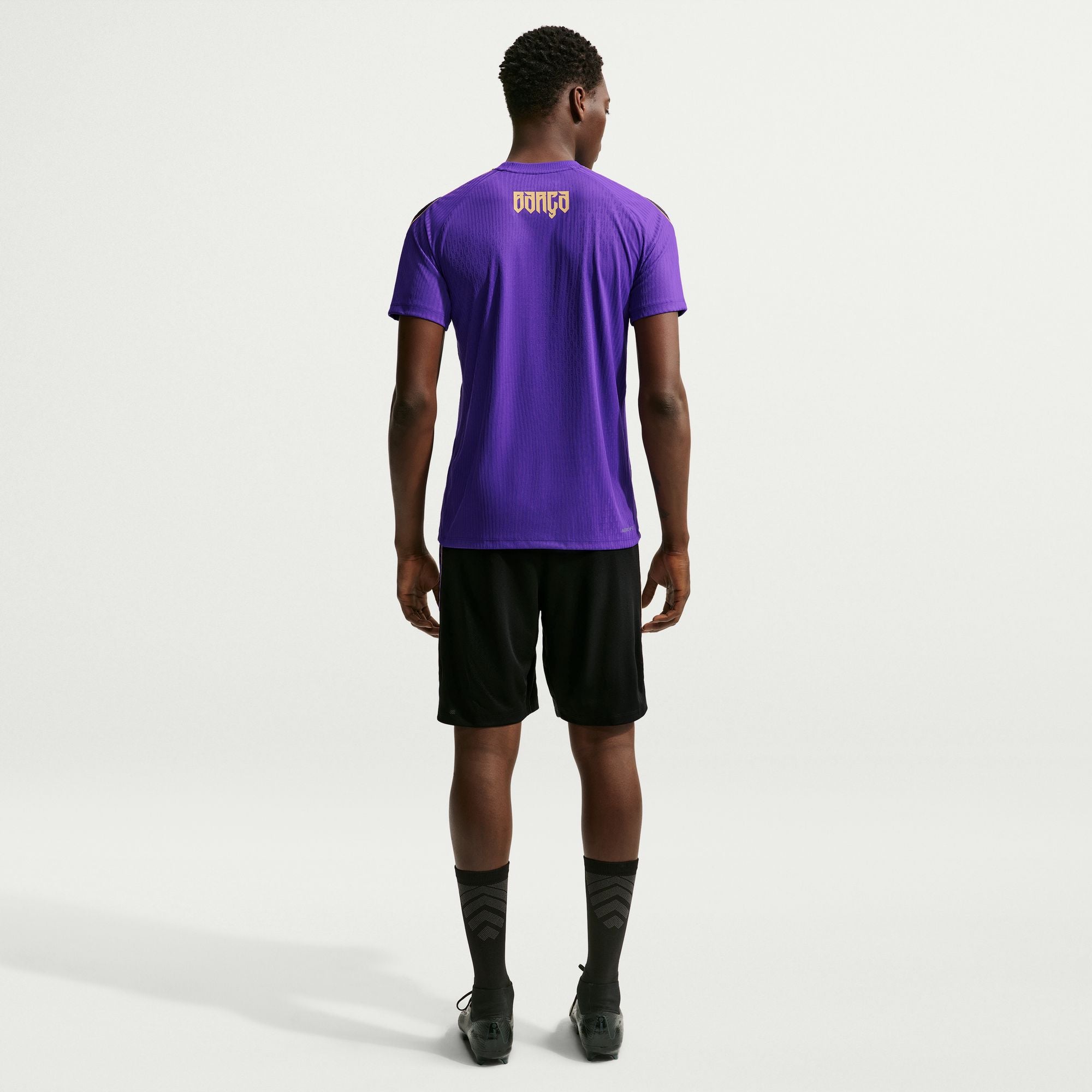 A man viewed from behind wearing a purple short-sleeve athletic t-shirt with a gold 'DONCA' logo on the upper back, paired with black athletic shorts, black socks, and black cleats, against a light gray studio background.